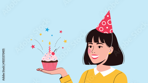 Happy young woman wearing a party hat holds a birthday cupcake with a lit candle and sprinkles.
