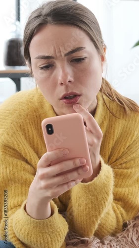 Confused young woman looking at smartphone screen, touching her lip and thinking about a problem or difficult decision while lying on sofa. Vertical video.
