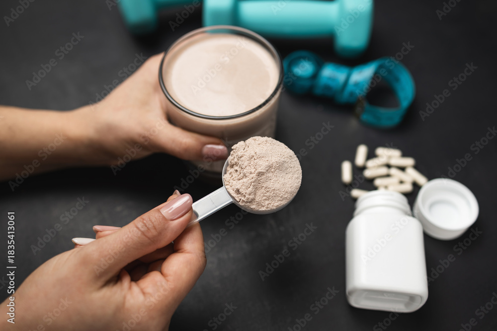 Fototapeta premium Female hand holding protein shake glass and scoop of chocolate whey protein powder on dark background with white capsules, dumbbells and measuring tape, concept of sport nutrition