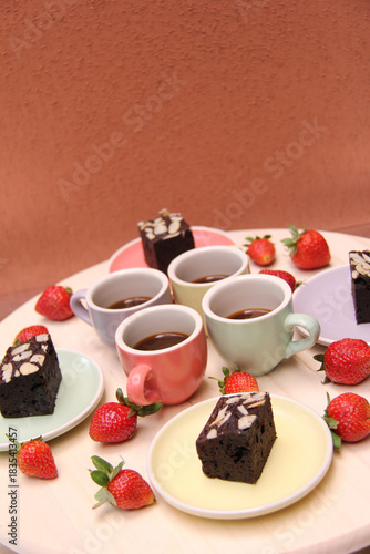 Vertical shot of a dessert and coffee setup featuring four espresso cups, almond brownies, and fresh strawberries on pastel plates against a warm textured background. Cozy café-style presentation.