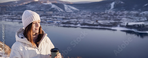 A Serene Winter Landscape featuring a Woman in a Cozy Sweater Sitting by a Tranquil River, A woman in a white jacket and hat drinks hot coffee in winter 