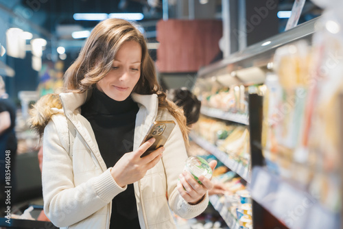 A smiling, beautiful blonde woman scans the label on a jar of yogurt, she studies the composition of the product, its properties and shelf life. Health and healthy food shopping,