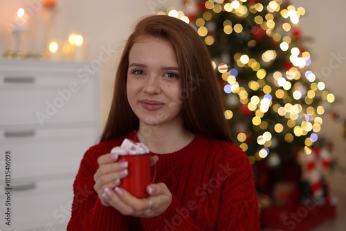 Beautiful teenage girl holding cup of hot drink with marshmallows against blurred lights at home. Christmas atmosphere