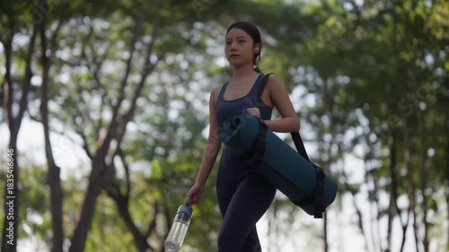 Young Asian woman carrying yoga mat water bottle walking through sunny park for morning fitness outdoor exercise wellness health active lifestyle journey