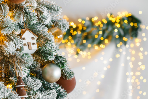Festive Christmas tree ornaments small house and round baubles on snowy spruce branch with bokeh illumination background with copy space. Holiday aesthetic decor for winter celebrations.
