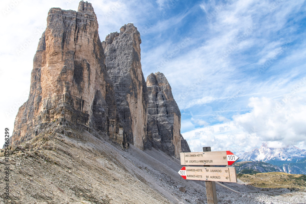Fototapeta premium Signage (destination location in Italian) of hiking trail 101 with north face of Tre Cime di Lavaredo peaks (Drei Zinnen) with white clouds in Dolomite Alps near Cortina d'Ampezzo, Italy