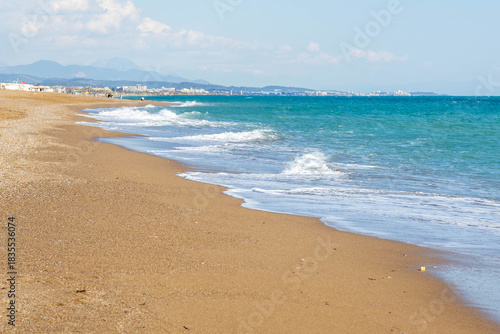 Fototapeta Naklejka Na Ścianę i Meble -  A serene empty beach with turquoise water and white foam on wet sand, mountains and hotels visible in the distance. A peaceful pre-season vista. Sorgun, Antalya, Turkey.