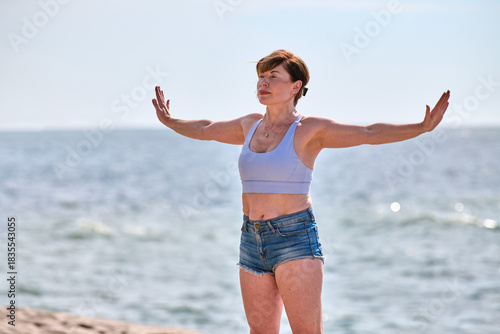 Young woman practicing yoga exercise with open arms by Baltic Sea shore in bright daylight