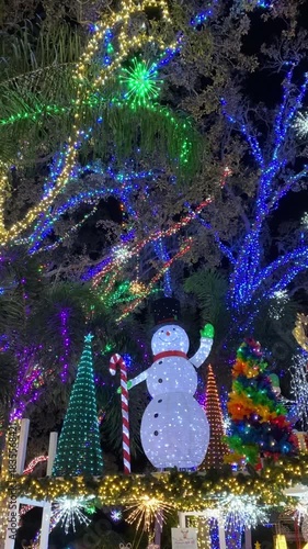 A giant Christmas tree with red bows glows under blue snowflake lights at night. A nutcracker, angel, and waving snowman stand among colorful LED decorations as people enjoy a festive park display.