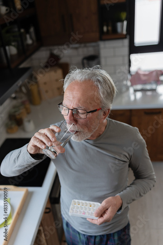 Elderly man in his kitchen taking daily medication, holding a pill and drinking water as part of his morning health routine