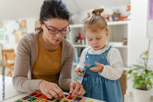 Smiling kindergarten teacher and toddler painting and crafting together at a bright classroom table, showing early learning, creativity, and child development in a warm preschool environment