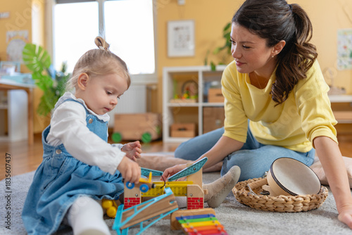 Kindergarten teacher and toddler sit on the floor playing with colorful wooden toys, encouraging early learning, creativity, and emotional bonding in a bright preschool classroom