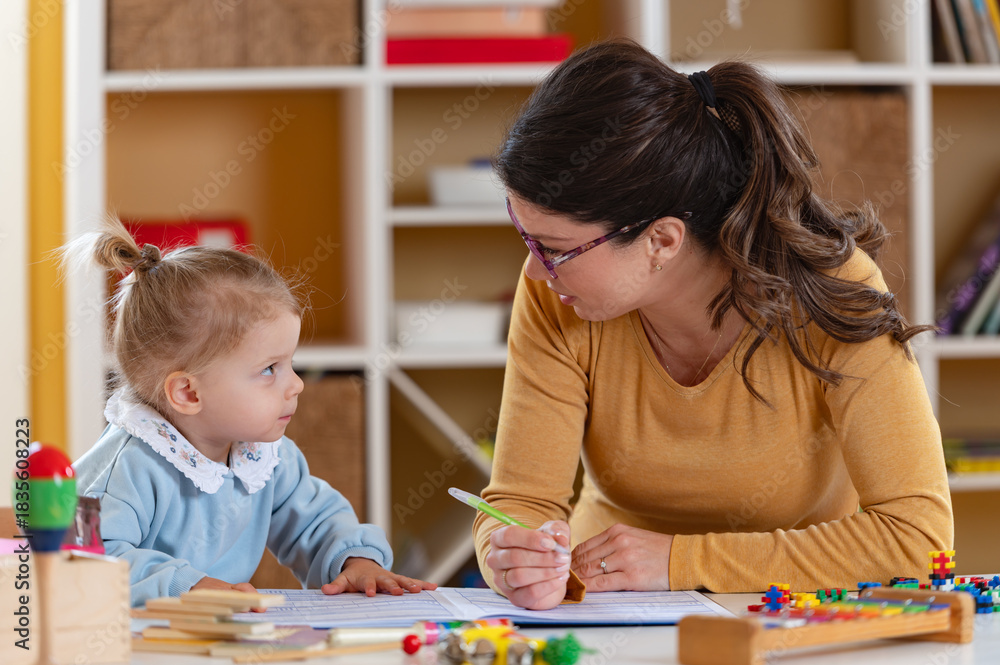 Fototapeta premium Adult woman guiding a toddler during a drawing activity at a table with colorful toys, supporting early learning and creativity in a bright home or classroom setting