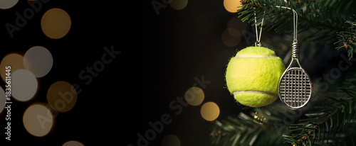 Tennis ball ornament and Christmas ornament racket, hanging from a spruce branch against a dark background with golden bokeh lights.