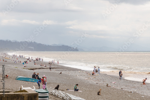 Kobuleti, Georgia - 21 August 2025: View of a pebbly beach with scattered people enjoying the day, the sea reflecting a muted sky, and distant mountains fading into the horizon.