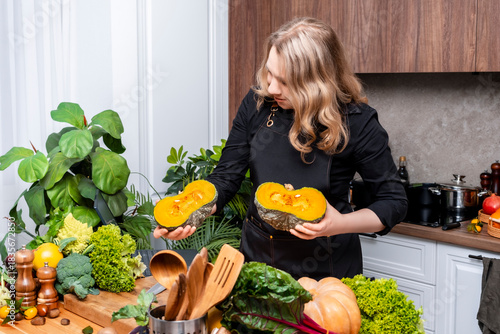Smiling young woman in black apron holding large green pumpkin on wooden kitchen table with fresh autumn vegetables and herbs during cozy seasonal home cooking