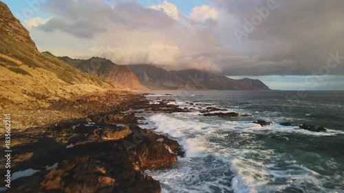 Kaena Point State Park. The rugged coastline of Hawaii with waves hitting the rocks. The sun sets behind mountains, casting warm light on the water and land.