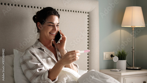 Obraz na plátne Smiling woman in cozy bedroom, holding pregnancy test while talking on smartphon