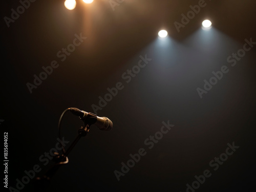 Single microphone on stand dramatically lit by stage spotlights against dark concert venue background.