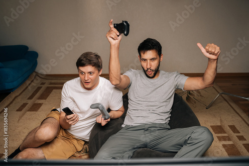 Obraz na plátne Happy excited two friends sitting on sofa cheer and raise controllers in celebration as they play video games together at home, smiling and enjoying competitive, casual gaming night
