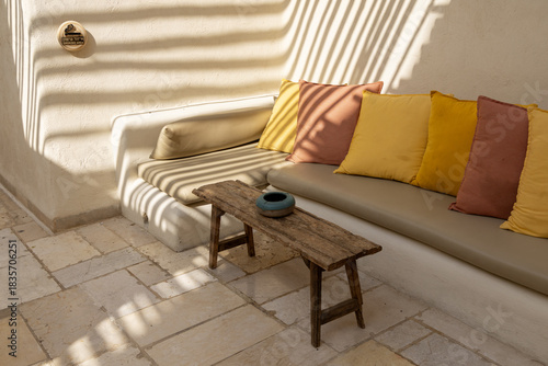 Inviting outdoor lounge with built in bench, soft cushions and colorful pillows under pergola slat shadows. Bright Mediterranean courtyard vibe ideal for hospitality lifestyle and design themes