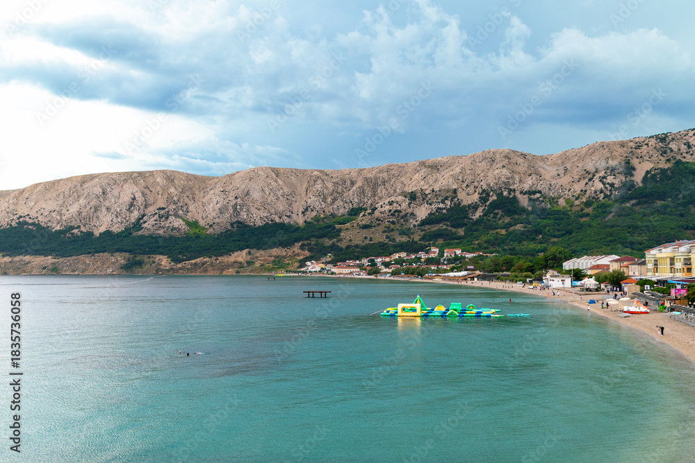 Obraz premium Scenic view of beach with inflatable water park and mountains under cloudy sky. Baška, Krk island, Croatia