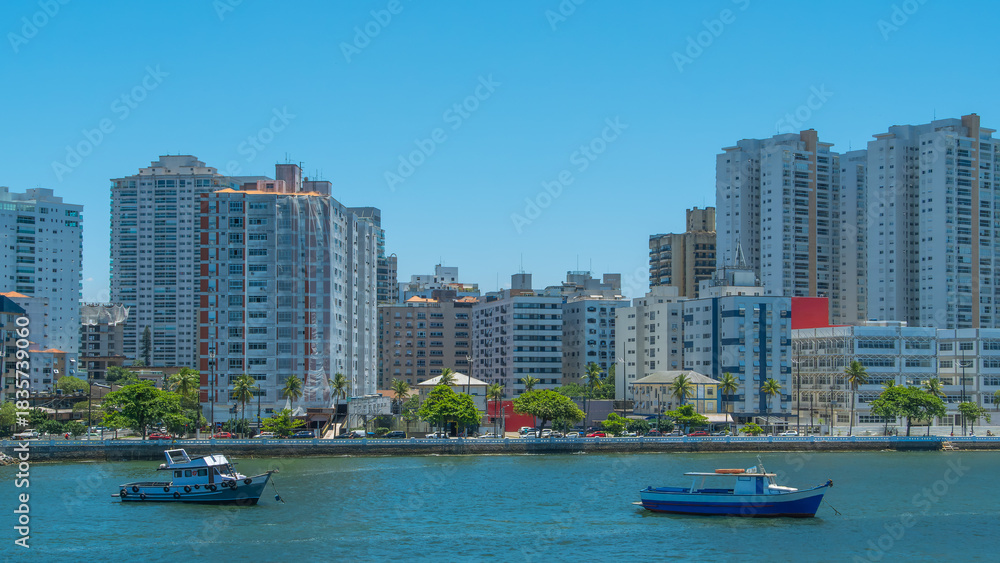 Fototapeta premium Bright, sunny view of a Brazilian city skyline, featuring modern apartment buildings overlooking a harbor with small fishing boats on the clear blue water