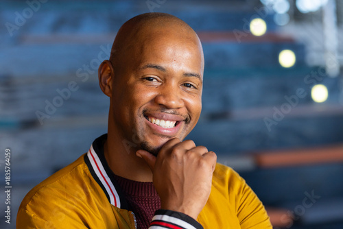 African American male smiling, resting chin in cafe lounge with string lights wearing bomber jacket
