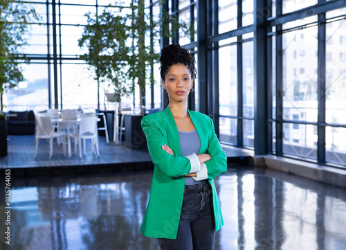 African American woman standing wearing green blazer and jeans in office lobby with potted plants