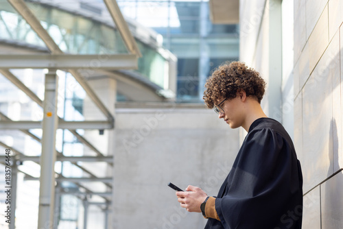Man wearing academic gown standing under glass canopy holding smartphone at walkway, copy space