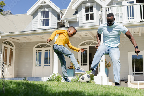 African American father and son kicking soccer ball on front yard wearing light-blue tee yellow top