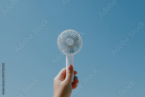 Handheld mini fan held up against clear blue sky symbolizing cool relief during hot summer days and portable convenience for outdoor activities