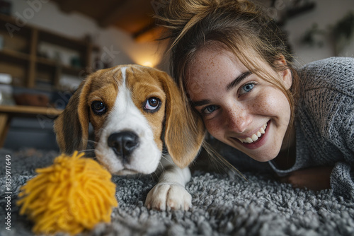 Young woman with freckles and blue eyes smiles happily while lying on carpet next to beagle puppy playing with yellow toy in cozy home setting