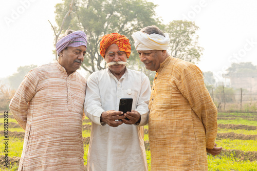 Three rural indian farmers using smart phone at agriculture field. Happy senior male friends check government scheme online on mobile phone. Wireless technology, Internet banking. Digital india