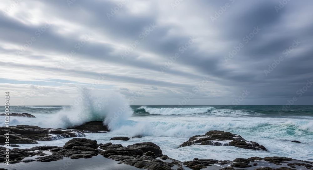 Obraz premium Turbulent ocean waves crash forcefully against dark coastal rocks beneath a sweeping cloudy sky