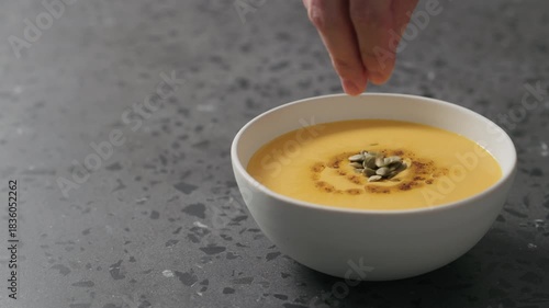 Slow motion pumpkin seeds add to pumpkin cream soup in white bowl on terrazzo countertop