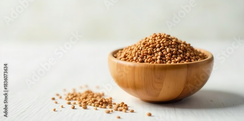 A heap of small, round, brown seeds piled in a natural wooden bowl with a scattering of seeds on a textured surface