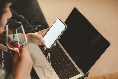 Woman holding smartphone with blank screen and drinking wine beside laptop, representing digital lifestyle,Businesswoman relaxation after work, social media browsing, or modern multitasking at home