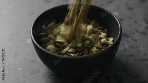 Slow motion dried pumpkin seeds falling into black bowl on terrazzo countertop