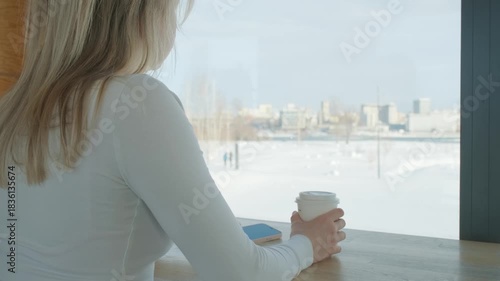 The caucasian young woman is drinking coffee in a coffee shop in winter in front of a window	