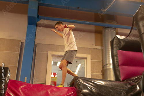 Teen boy balancing on inflatable obstacle showing playful confidence and dynamic motion in indoor softplay gym white shirt and shorts, arms outstretched for balance, vibrant cushions and padded