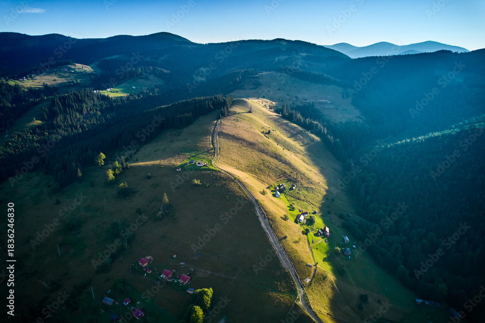 Obraz premium Aerial view of picturesque valley with rolling hills, lush forests, and scattered houses. Sunlight bathes landscape in warm glow, highlighting vibrant greens and creating serene atmosphere.