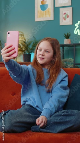Happy young child kid girl blogger communicating video call online waving with social media subscribers, taking selfie on smartphone. Female teen recording video story at home in room sits on sofa.