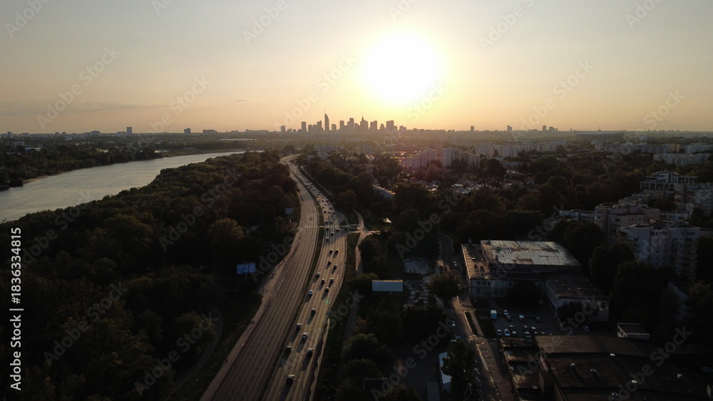 Obraz premium Smooth drone footage flying toward the Warsaw city skyline during a warm summer sunset. The video captures the Vistula River, extensive green areas, residential neighborhoods, and a major highway fil