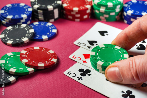 A player is focused on their hand in a poker game, placing a green chips next to three fives showing on the table. Colorful chips surround the cards, creating a lively casino atmosphere