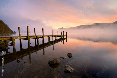 Misty Derwentwater sunrise with wooden jetty leading out over calm water. Lake District, England, UK.