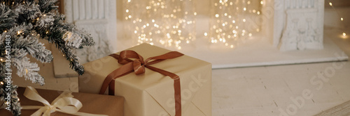 Two wrapped Christmas presents with ribbons under snowy tree near fireplace in cozy room with warm fairy lights and white wooden floor. Concept: holiday, winter, celebration, home decor.