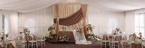 Elegant wedding banquet hall with draped ceiling, chandeliers, floral arrangements, and decorated stage in beige and white tones. Concept for wedding venue, celebration, luxury event, decor design.