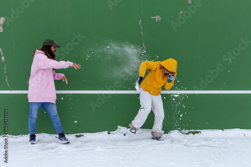 Two people playing in the snow against a green wall, throwing snowballs in a fun winter scene. Dynamic outdoor action capturing joy, movement, cold weather and playful seasonal activity