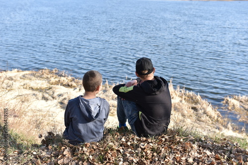 Tablou pe pânză Two boys sitting on a grassy riverbank and looking at the calm water on a sunny day